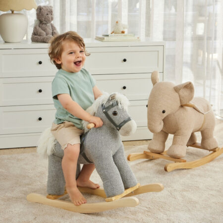Little boy toddler laughing while riding on small grey tweed rocking horse with a tweed rocking elephant in the background of the nursery.